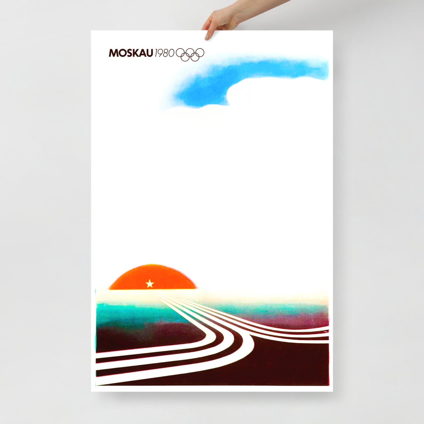 A white skinned hand holds large Moscow 1980 Olympic Games poster with text MOSCAU, Olympic logo and minimalist composition with rising sun and roads made of Moscow Olympics logo going towards it. From artsoviet.com
