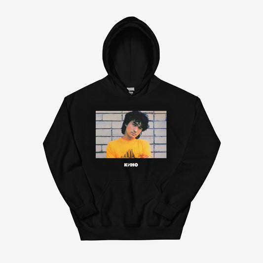 Rare archival photo of Viktor Tsoi in a yellow T-shirt against a white brick wall with a little KINO band logo under the photo, printed on the front of a heavyweight quality black unisex hoodie with a KINO logo on the back neck side, from GruppaKino.com. Front side.